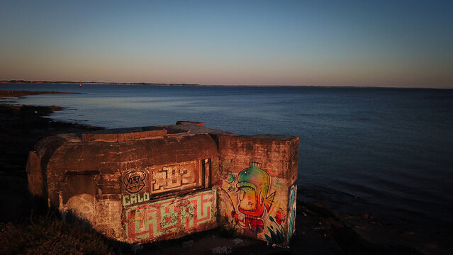 Aerial view of a WW2 Atlantic Wall bunker covered in colorful graffiti situated on the rocky coastline at sunset with the calm sea in the background in Damgan, Bretagne, France.