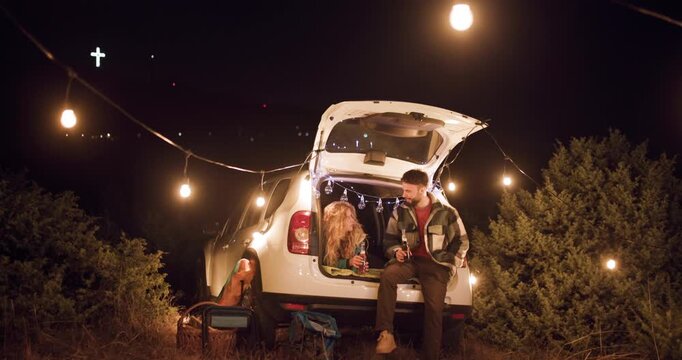 Enjoying a night under the stars with friends at a campsite in the hills with a car and string lights at sunset