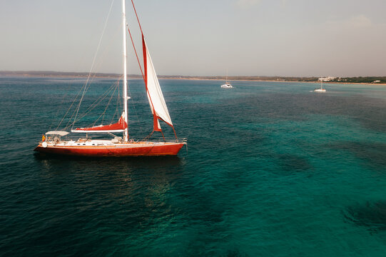 Aerial view of a red sailboat with white sails anchored in the clear turquoise waters off the coast of Formentera, Balearic Islands, Spain.