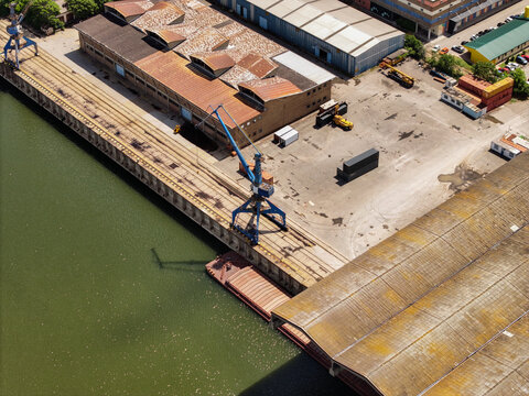 Aerial view of the Mini skladista Luka Beograd port facilities with industrial warehouses, a blue crane, and a barge docked along the river bank in Beograd, Grad Beograd, Serbia.