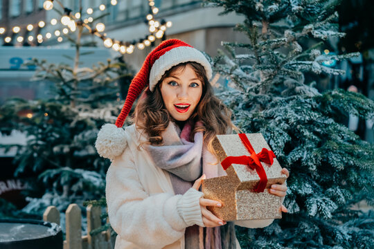 Happy surprised woman opening Christmas gift box on outdoor Xmas fair and New Year celebration on Christmas market. Festive surprise winter miracle. Wow emotions,happiness and winter holidays concept