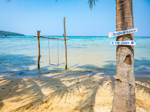 Alano Beach in Karimunjawa, Indonesia, featuring a wooden swing in shallow turquoise water next to a palm tree with Bikini and Beach and Tan Lines signs, creating a serene tropical travel destination