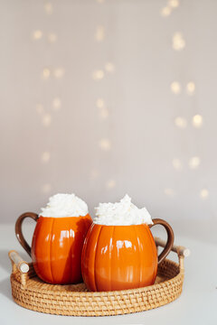 Pumpkin shaped cups of hot drink with whipped cream on woven tray with blurred garland background. Pumpkin latte with spices. Cozy autumn beverage for Thanksgiving or Halloween holidays at home