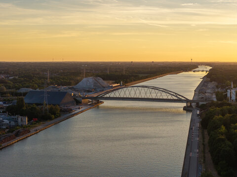 Aerial view of the Albert Canal featuring a modern arched bridge, industrial facilities, and wind turbines under a golden sunset sky in Genk, Flanders, Belgium.