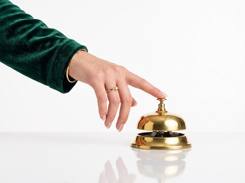 Hand in green velvet sleeve pressing golden hotel reception bell isolated on solid white background