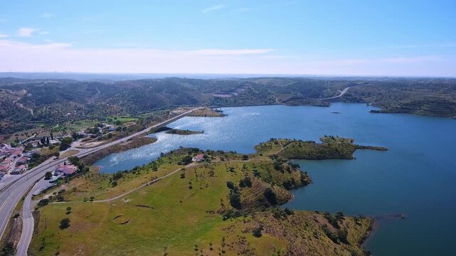 Sweeping aerial view of the odeleite dam and its blue water reservoir, surrounded by green hills and a small village in the algarve region of portugal under a clear summer sky