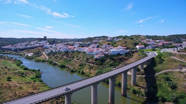 Aerial view tracking a lone car crossing a long concrete bridge over the guadiana river, with the scenic historic town of mertola and its castle visible in the sunny alentejo landscape
