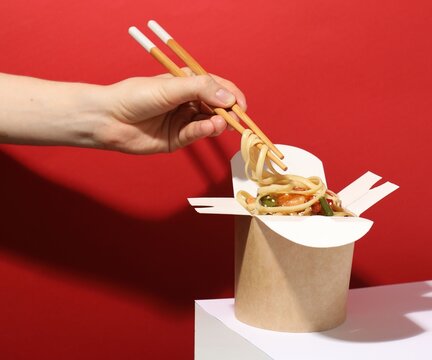 Woman eating tasty takeaway wok with chopsticks at white table on red background, closeup