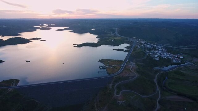 Majestic aerial view of the odeleite dam and reservoir in portugal, showing the tranquil water reflecting the warm colors of the setting sun over the beautiful algarve landscape and nearby village