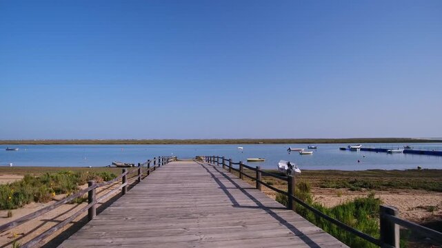 Point of view walking along a long wooden pier towards the calm waters of the ria formosa lagoon in fuzeta, algarve, portugal, on a bright sunny day with a clear blue sky