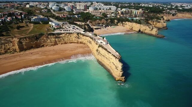 Dramatic aerial footage revealing the iconic chapel of our lady of the rock perched on a stunning cliff promontory, surrounded by turquoise ocean waters and sandy beaches in algarve, portugal