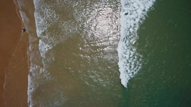 Incredible aerial top down shot showing the majestic ocean with its powerful waves crashing on the shore and surfers enjoying the water, creating a stunning and relaxing seascape background