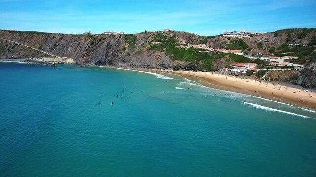 Majestic aerial view of surfers catching waves in the turquoise ocean at arrifana beach, a popular holiday destination with scenic cliffs and golden sand in the sunny algarve region, portugal