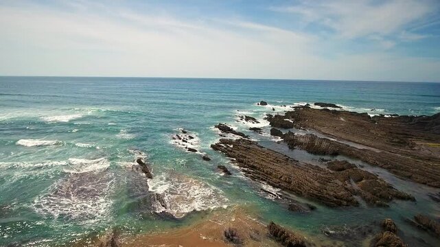 Stunning aerial footage of the wild atlantic ocean with turquoise waves breaking on the rugged cliffs and dramatic rock formations of costa vicentina in aljezur, portugal during a sunny day