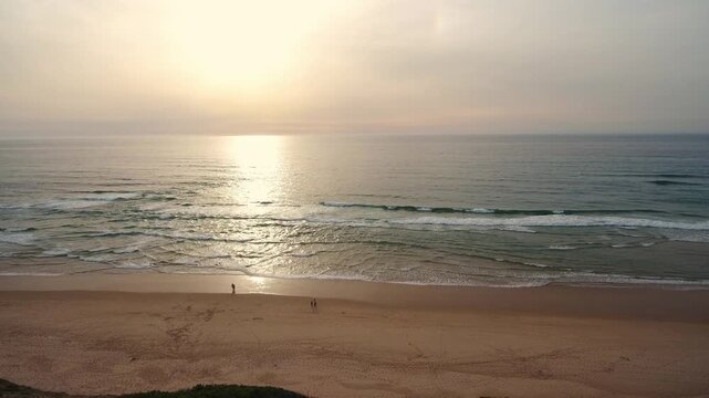 Aerial view of a sandy beach with people walking along the shore as the sun sets over the ocean, its golden light reflecting on the gentle waves creating a peaceful and serene atmosphere