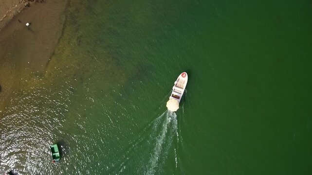 Aerial top down view showing a small white motorboat sailing across the beautiful green sea, leaving a trail of wake on the calm water surface during a sunny summer day on the coast