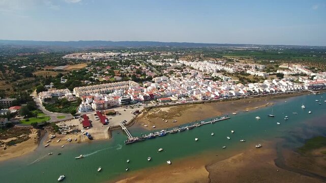 Stunning aerial perspective flying over the coastal town of cabanas de tavira in the algarve region, showing the waterfront, boats on the water, and traditional white buildings