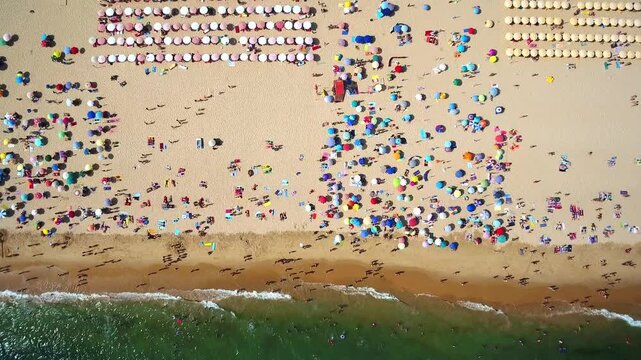 Bird's eye view of a bustling sandy beach filled with many colorful umbrellas and people sunbathing, swimming in the turquoise ocean, and enjoying their summer vacation on a sunny day