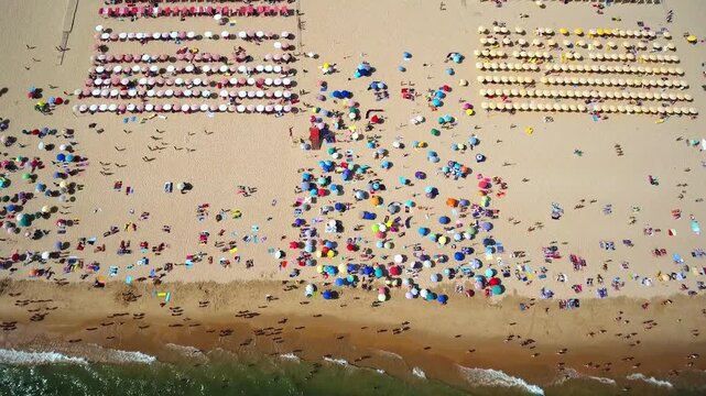 Crowds of people relaxing and swimming on a beautiful sandy beach with colorful umbrellas during a sunny summer day, showcasing a popular travel destination and vacation hotspot from above