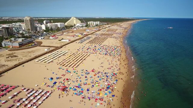 Thousands of people enjoying a sunny summer day on the crowded sandy beach of monte gordo in algarve, portugal, with colorful umbrellas dotting the coastline next to the turquoise sea