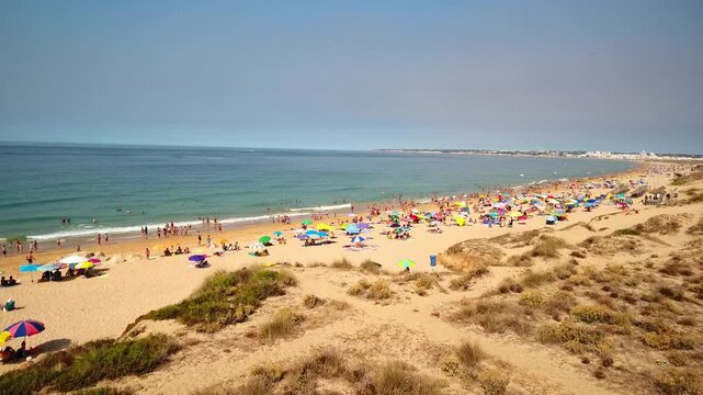 Aerial view flying over sand dunes revealing a crowded beach with many people sunbathing under colorful umbrellas and swimming in the atlantic ocean during a sunny summer day in algarve, portugal