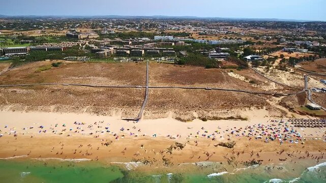 Aerial view flying backwards over a crowded sandy beach with tourists enjoying the summer sun and swimming in the beautiful turquoise ocean waters of the atlantic coast in portugal