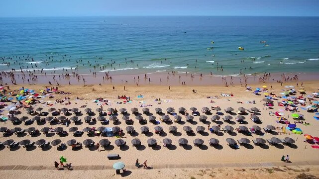 Sunny day with many tourists relaxing on the sand under umbrellas and swimming in the beautiful blue ocean, showcasing a popular destination during the peak of the holiday season