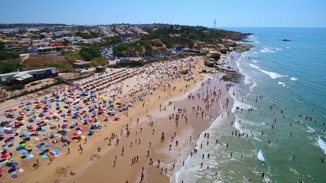 Aerial drone view of praia da gale coastline in algarve, portugal, sunbathers and colorful umbrellas on golden sand, swimmers in the atlantic on a busy summer day