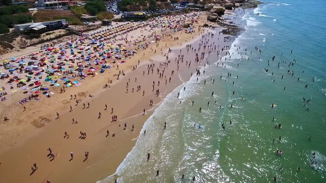 Sunny day aerial shot of the crowded gale beach in albufeira, portugal, with numerous tourists relaxing under colorful umbrellas, swimming in the ocean, and enjoying their summer vacation