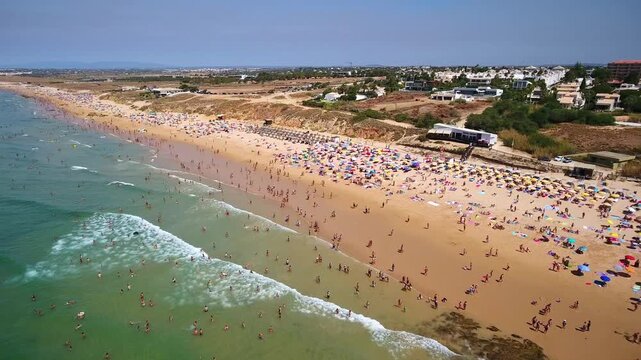 Aerial view of the long, sandy gale beach in albufeira, algarve region, with numerous tourists swimming in the atlantic ocean and relaxing under colorful umbrellas during a sunny day