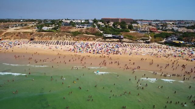 Aerial view of many tourists swimming in the clear ocean and relaxing under colorful umbrellas on the sandy shore of gale beach in albufeira on a hot summer day in algarve, portugal