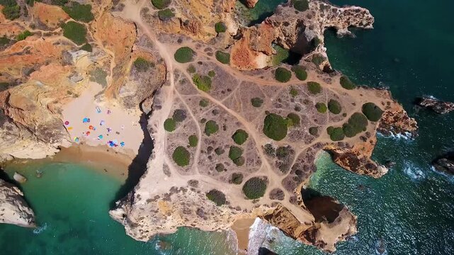 Majestic aerial top down view of people with colorful umbrellas relaxing on a secluded sandy beach surrounded by imposing golden cliffs and turquoise waters in algarve, portugal
