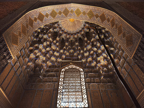 Ornate muqarnas ceiling inside Gur-e-Amir mausoleum in Samarkand with gilded honeycomb vaulting and geometric lattice window. Ideal for travel publications, cultural heritage features, architecture.