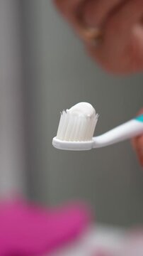 Close-up of white toothpaste being applied from a tube onto a white toothbrush, with a hand holding both items against a softly blurred background.