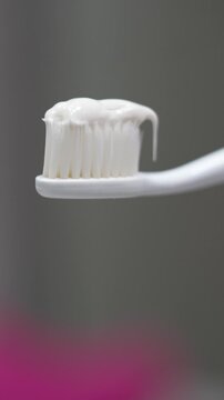 Extreme close-up of a white toothpaste tube dispensing toothpaste onto the bristles of a white toothbrush with a turquoise handle, set against a blurred background.