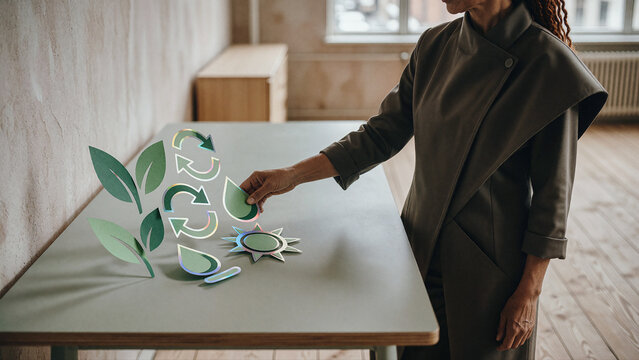 Woman reaches toward green paper cutouts of leaves and arrows arranged on a pale grey desk in a bright room with natural sunlight casting soft shadows.
