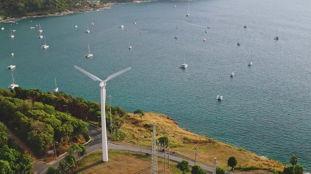 Wind turbine blades spinning, creating renewable energy on lush green hillside overlooking tranquil bay filled with many anchored sailboats and the inviting turquoise ocean. Windmill Viewpoint, Phuket