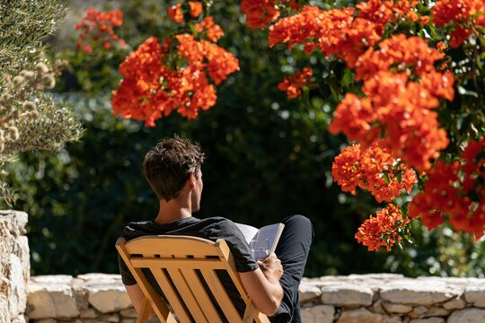 Man reading outdoors in a garden chair by bright orange flowers for relaxation background and summer leisure poster