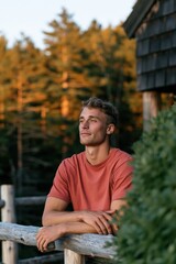 Fototapeta premium Young man leaning on rustic railing at golden hour with pine forest view for outdoor lifestyle background and wellness poster