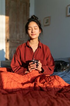 Young woman meditating with cup in bed in morning sunlight on cozy red bedding for calm bedroom background and relaxation design