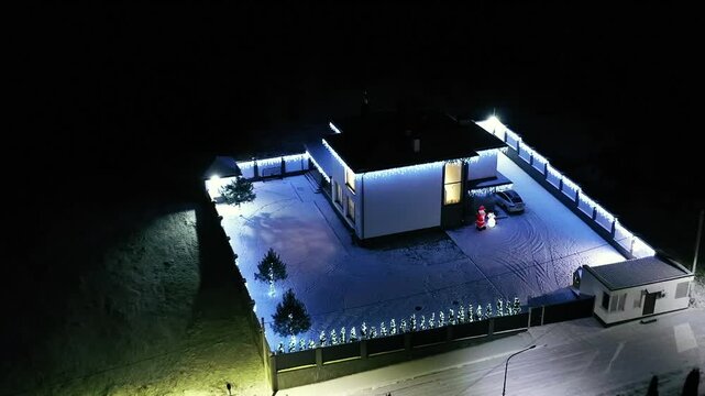 Snow blankets modern house at night. Lights outline the property's edges and roof. Single door glows warmly from within. No people are visible. Ideal for holiday, winter, or modern architecture themes