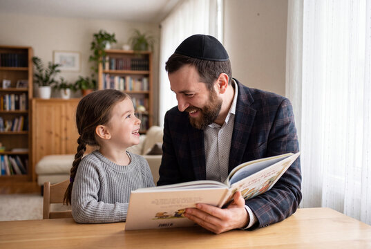 Jewish father wearing a kippah or yarmulke reading a storybook with his smiling young daughter at home.