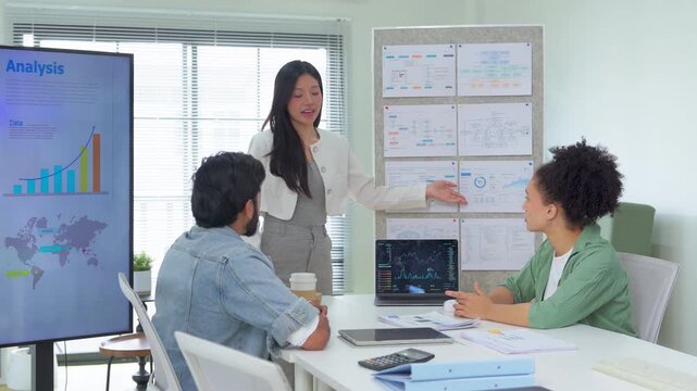 A presenter stands before a donut chart labeled "Marketing Research," addressing two seated colleagues with laptops and documents on the table a professional scene of marketing data presentation	