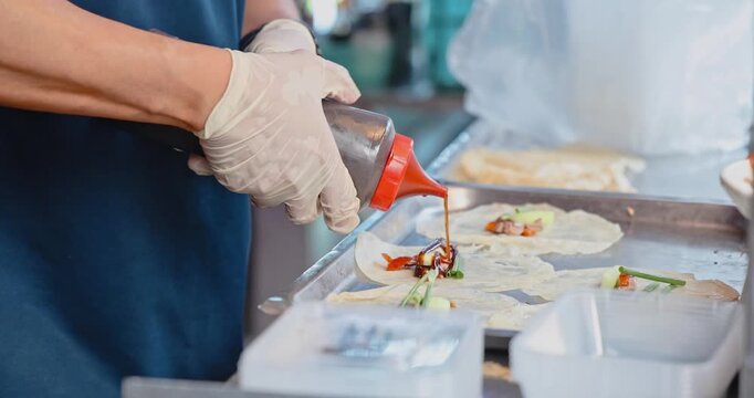 Chef hands in gloves squeezing dark sauce onto wraps, commercial kitchen food preparation, Asian street food stall, professional culinary service.