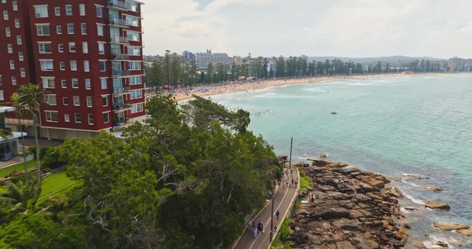 Vibrant Sydney Manly Beach coastline with people enjoying scenic walkway and calm ocean waters, framed by residential buildings and lush green vegetation under a cloudy sky. Aerial view drone flight