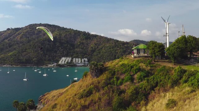 Windmill Viewpoint, paraglider soaring above the tropical coastline of Nai Harn Beach, Phuket, Thailand. Anchoring boats in the bay, green hills, a viewpoint gazebo, and a wind turbine. Aerial shot