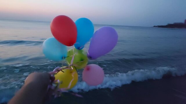 Woman holding colorful balloons in hand flying in wind over sea. Bunch colorful inflatable balloons on background of sea at dawn and sunset. Person playing multicolored air balloons. Concept fun