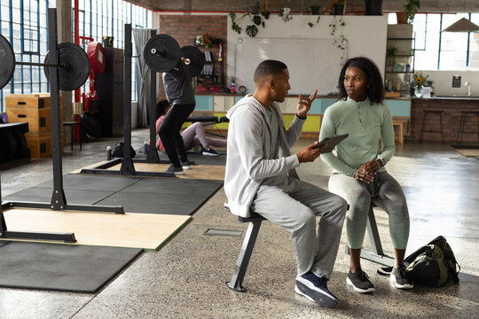 African American coach guiding woman in sportswear on bench at gym, holding tablet, duffel nearby