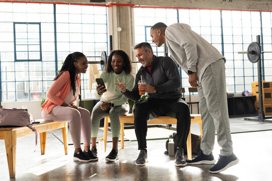 Diverse friends sitting on wooden benches at gym in athletic wear, sharing smartphone