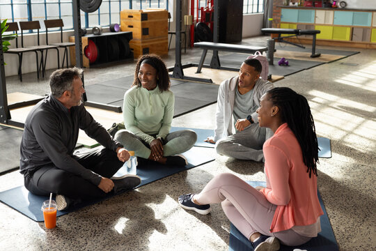 Diverse friends sitting on exercise mats chatting between sets in bright gym with weights